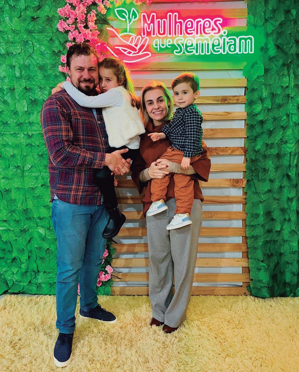 Liana Faccio Iaguszeski com o esposo Marcos e os filhos, em frente a um painel do evento Mulheres que Semeiam