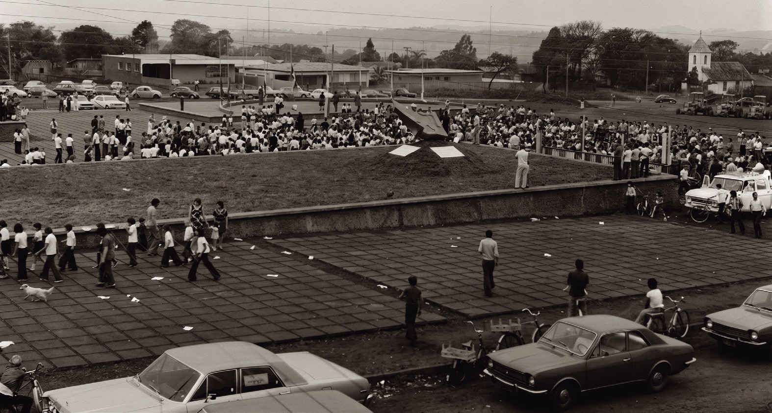 Multid&atilde;o reunida em pra&ccedil;a com autom&oacute;veis estacionados ao redor, em foto de 1976