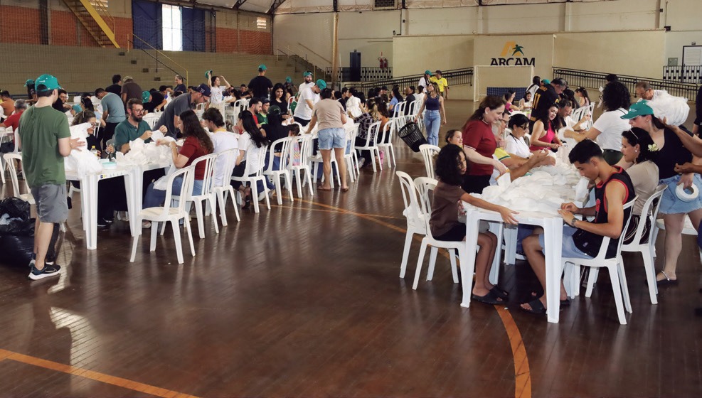 Funcion&aacute;rios e familiares sentados em mesas no gin&aacute;sio, confeccionando fraldas