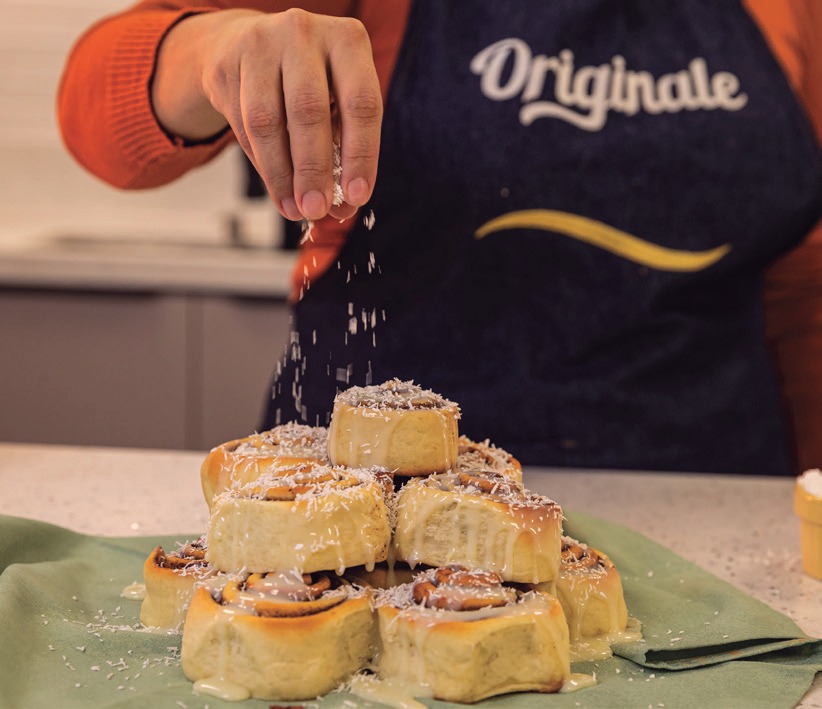 M&atilde;o polvilhando coco ralado sobre uma pilha de cinnamon rolls rec&eacute;m-assados